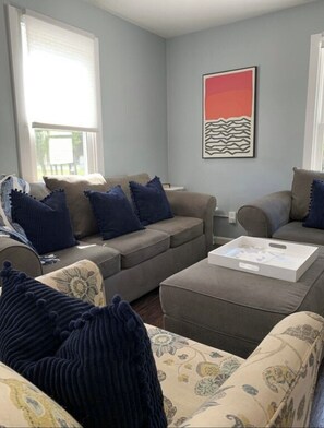 Un salon avec un ensemble de canapés gris, des coussins bleus et beige, une table basse blanche et un tableau abstrait encadré.