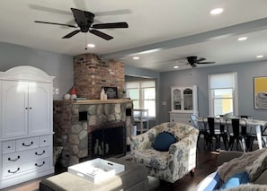 A living room with a stone fireplace, white cabinetry, a patterned armchair, and a ceiling fan.