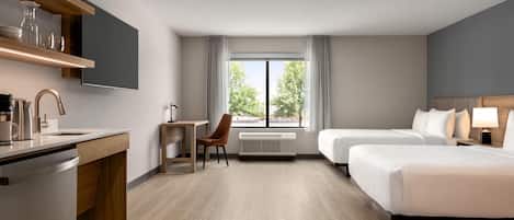 Habitación de hotel con kitchenette, escritorio, dos camas, una silla y un gran ventanal con vistas a árboles.