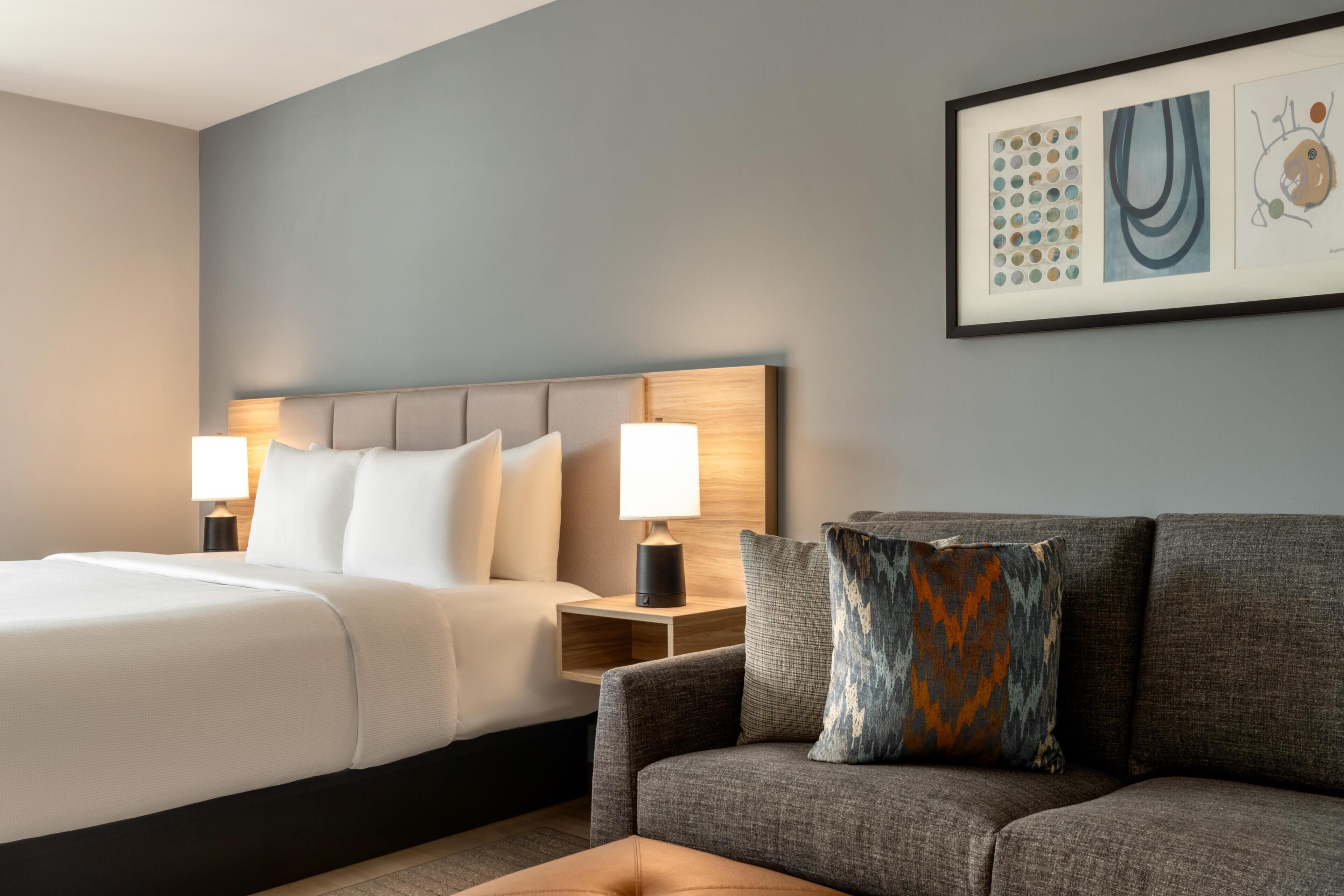 A hotel room with a bed, a nightstand with a lamp, a sofa, and a wall with framed artwork.