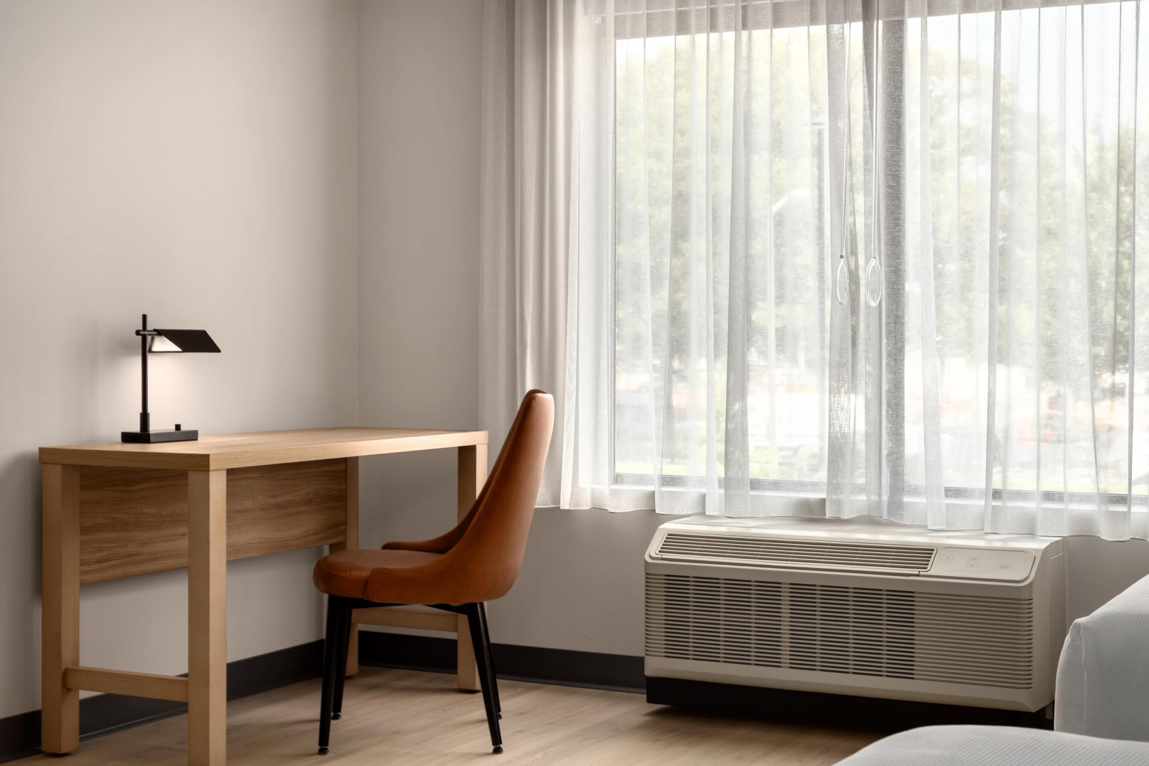 A room with a wooden desk, a brown chair, a white air conditioner, and a window with sheer curtains.