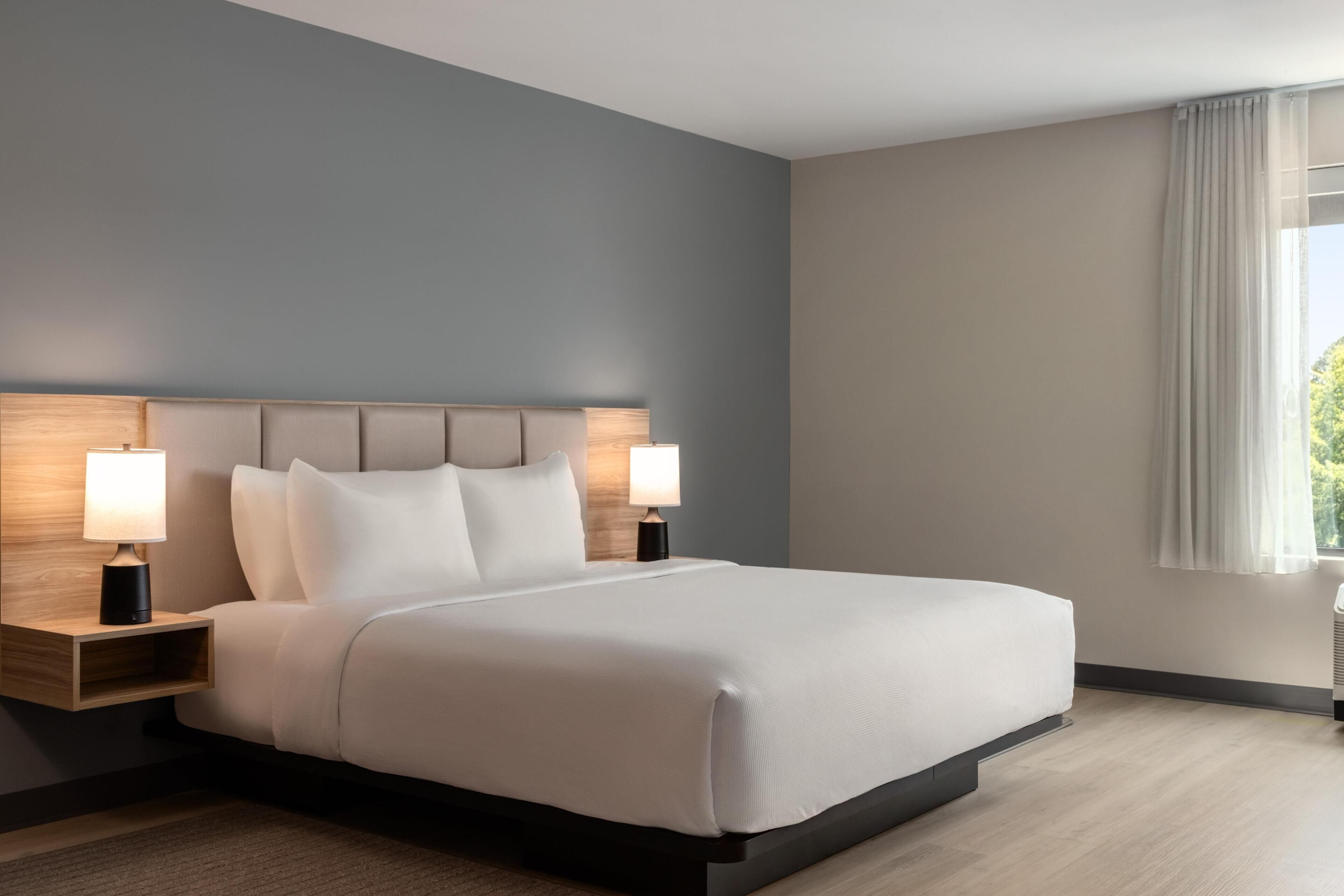 Habitación de hotel moderna con una cama grande, dos mesitas de noche con lámparas, un cabecero de madera y vistas a zonas verdes por la ventana.