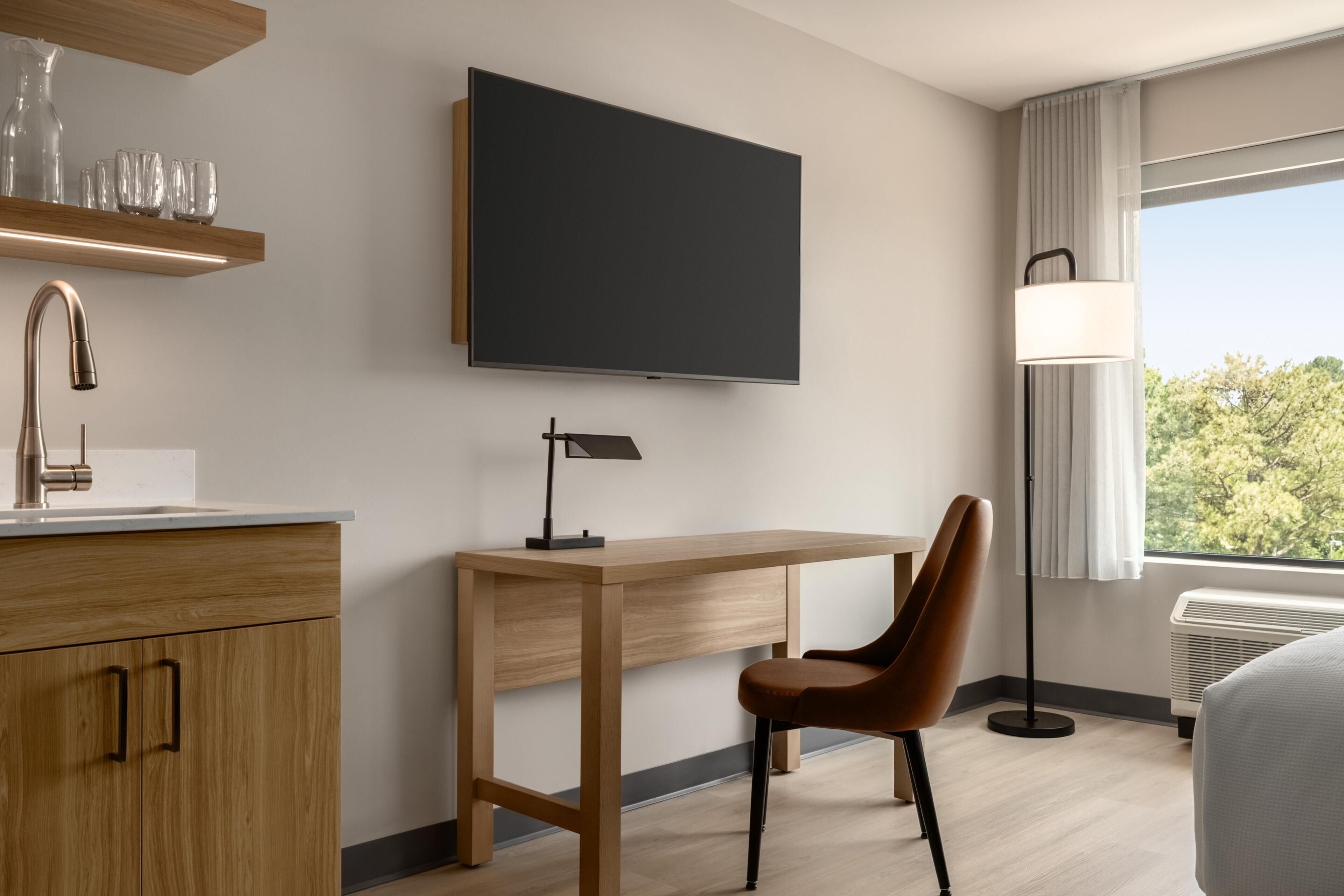 A hotel room with a wooden desk, a chair, a flat-screen TV, a modern sink, and a window with a view of trees.