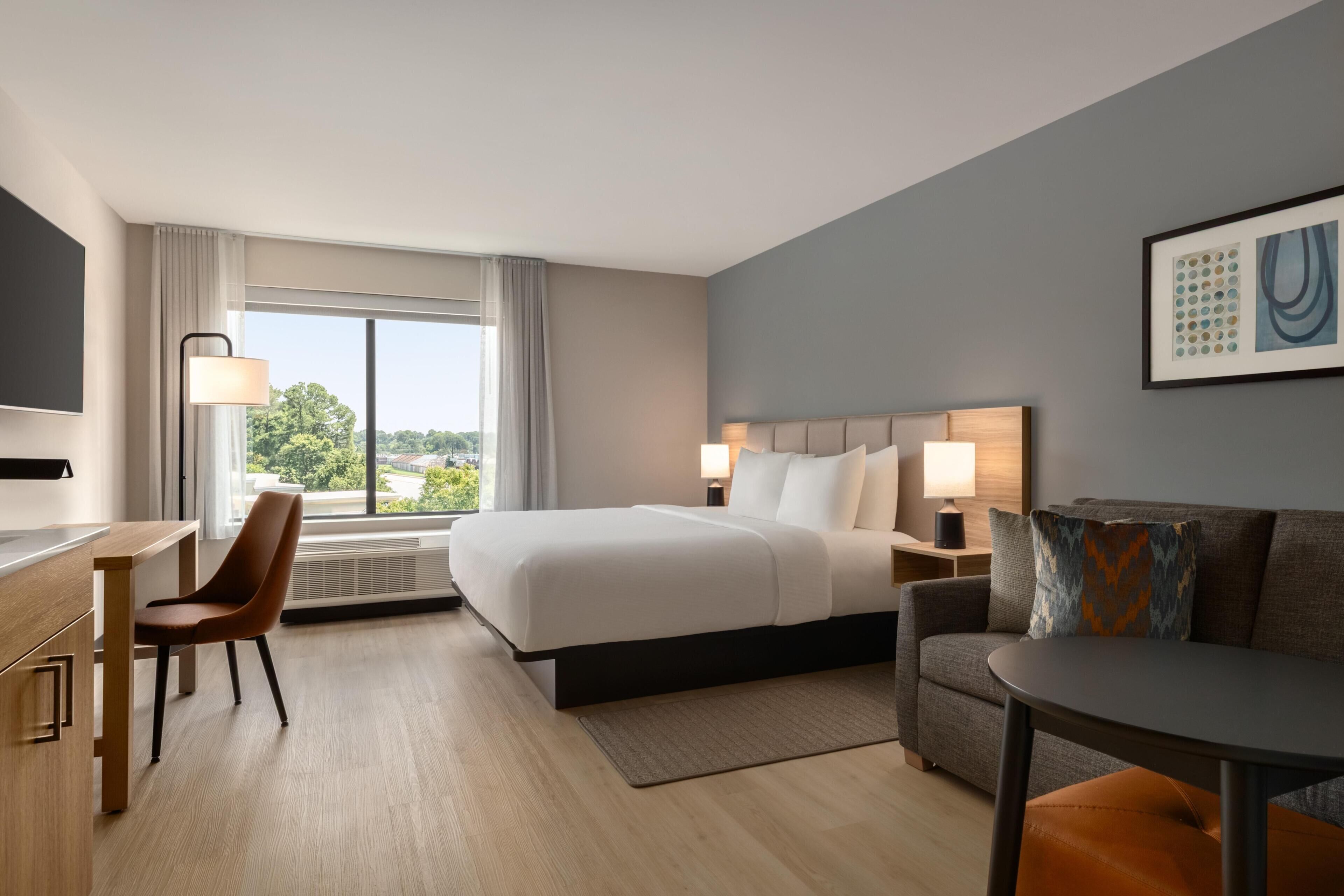 A hotel room with a bed, a desk, a chair, a sofa, and a large window offering a view of the outdoors.