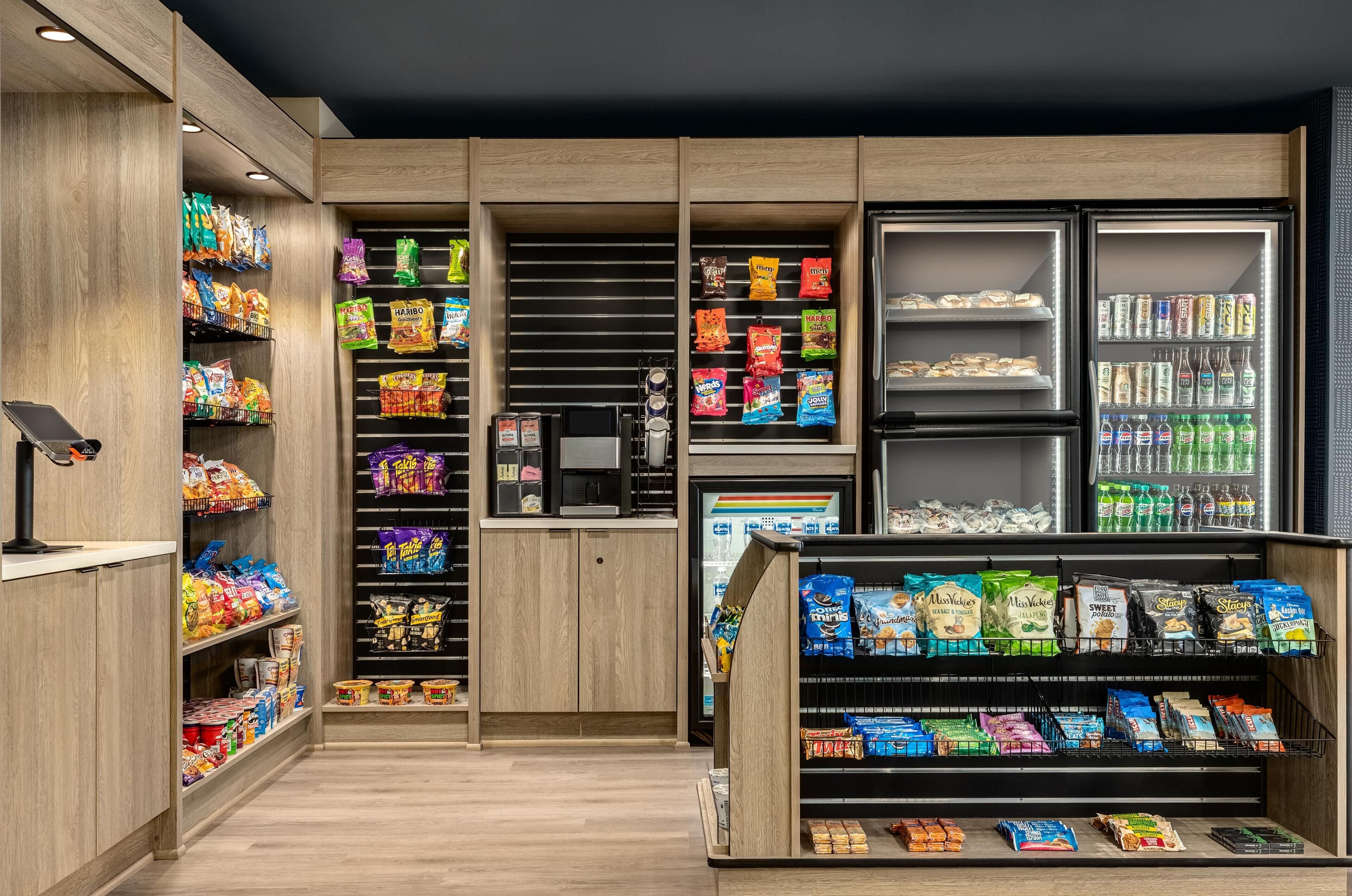 Zona de exhibición de aperitivos y bebidas en una tienda, con diversos productos empacados, una máquina de café y bebidas refrigeradas.