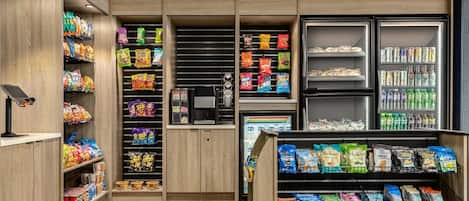 Zona de exhibición de aperitivos y bebidas en una tienda, con diversos productos empacados, una máquina de café y bebidas refrigeradas.