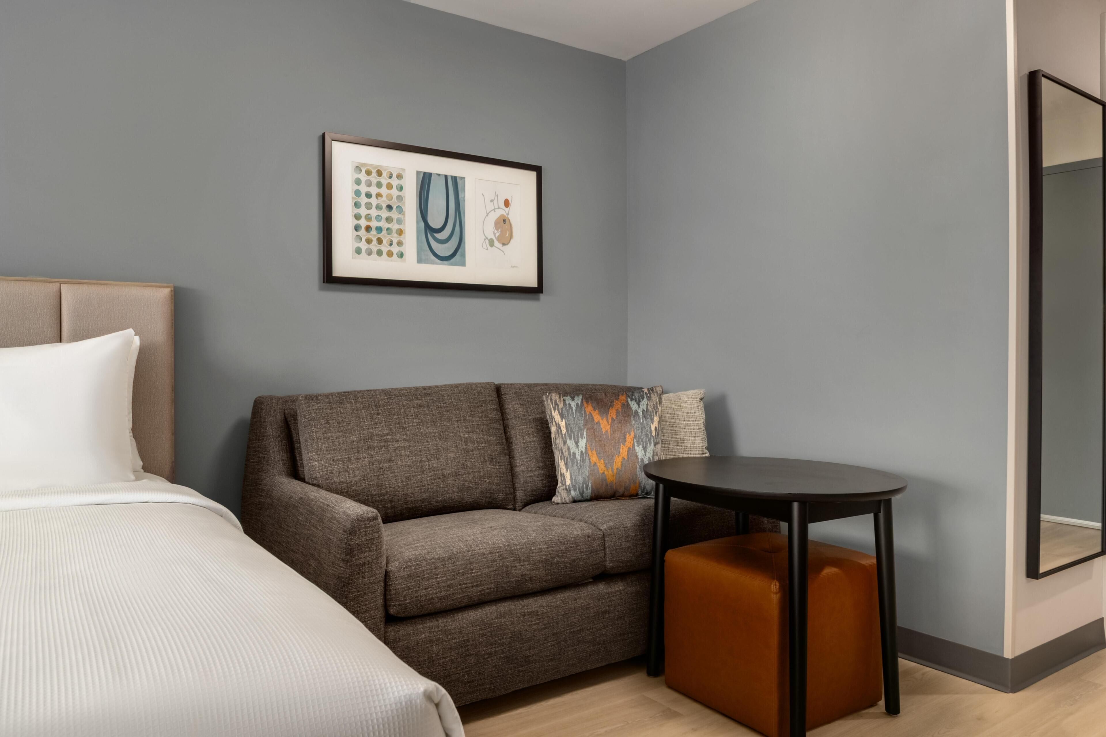 A hotel room with a bed, a sofa, a small table, and a framed artwork on the wall.