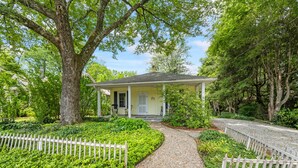 Una casa de un piso con un porche delantero, rodeada por un camino de grava y una cerca blanca de piquetes.