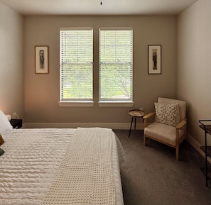 Ein Schlafzimmer mit einem Bett, zwei Fenstern mit Jalousien, einem Sessel, einem kleinen Tisch und zwei gerahmten Bildern an der Wand.