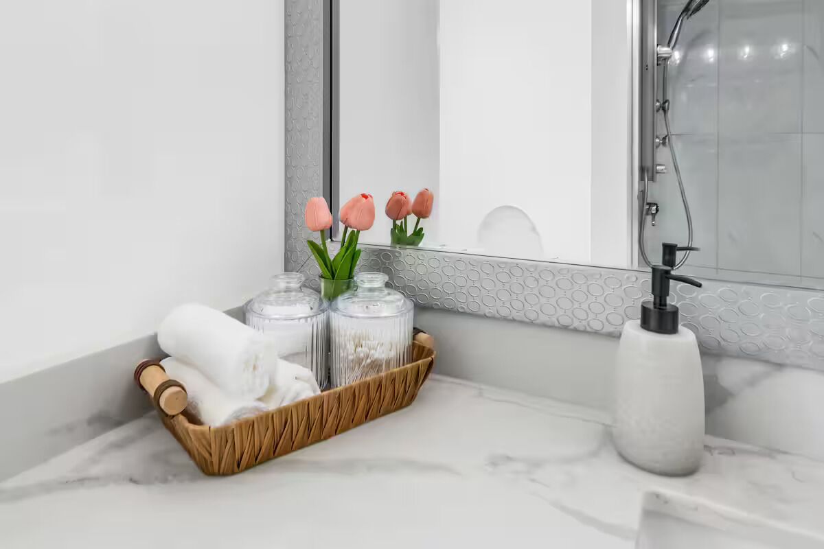 Ein Badezimmer mit Marmorplatte, einem Holztablett mit Toilettenartikeln, einer Glasvase mit Blumen, einer weißen Seifenspender und einem Spiegel.