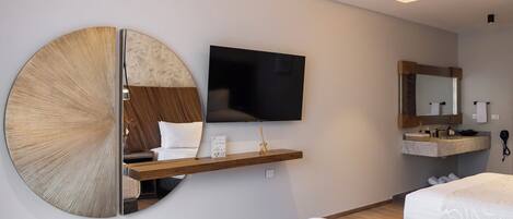 A modern hotel room with a large wall-mounted TV, a round mirror, a wooden shelf with a small table, a bed with patterned bedding, and a bathroom with a vanity and mirror.
