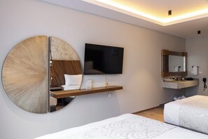 A modern hotel room with a large wall-mounted TV, a round mirror, a wooden shelf with a small table, a bed with patterned bedding, and a bathroom with a vanity and mirror. - HOTEL BOUTIQUE ALMA (Jiutepec)