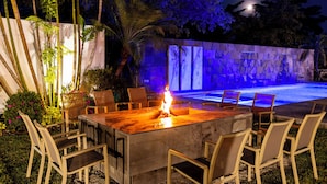Un área de comedor al aire libre con una chimenea, rodeada de sillas y una piscina iluminada en azul.