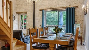 A dining area with a wooden table set for four, surrounded by chairs with cushions. A staircase is visible to the left, and a window with curtains is in the background. - Gîte Les Magnolias Logis Bourseguin, private terrace and Wi-Fi (Bourneau)