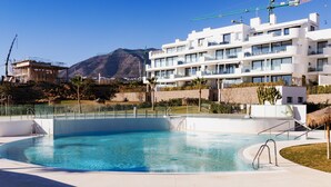Una piscina con agua azul cristalina, rodeada por una solana de hormigón, y un edificio moderno con balcones al fondo.