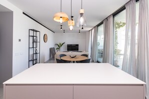 A modern kitchen with a large island, pendant lights, a dining area with a round table, and a TV in the background. - Cozy apartment with a large terrace, nice views, AC in a peaceful community (AN)