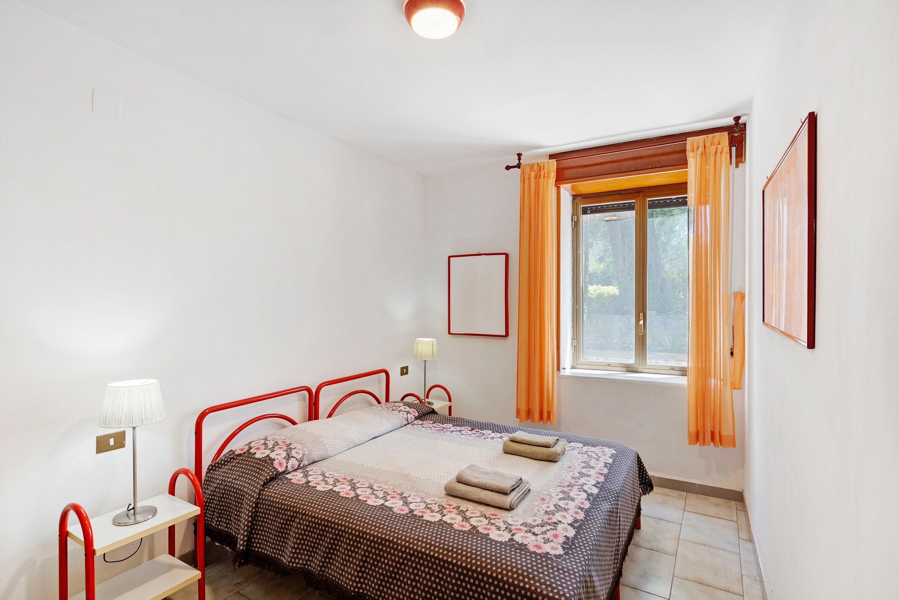 Ein Schlafzimmer mit Bett, Nachttisch, Lampe, Fenster mit Vorhängen und einem an der Wand hängenden Bilderrahmen.