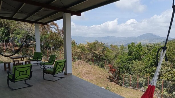 A covered patio with green chairs and a hammock, overlooking a mountainous landscape.