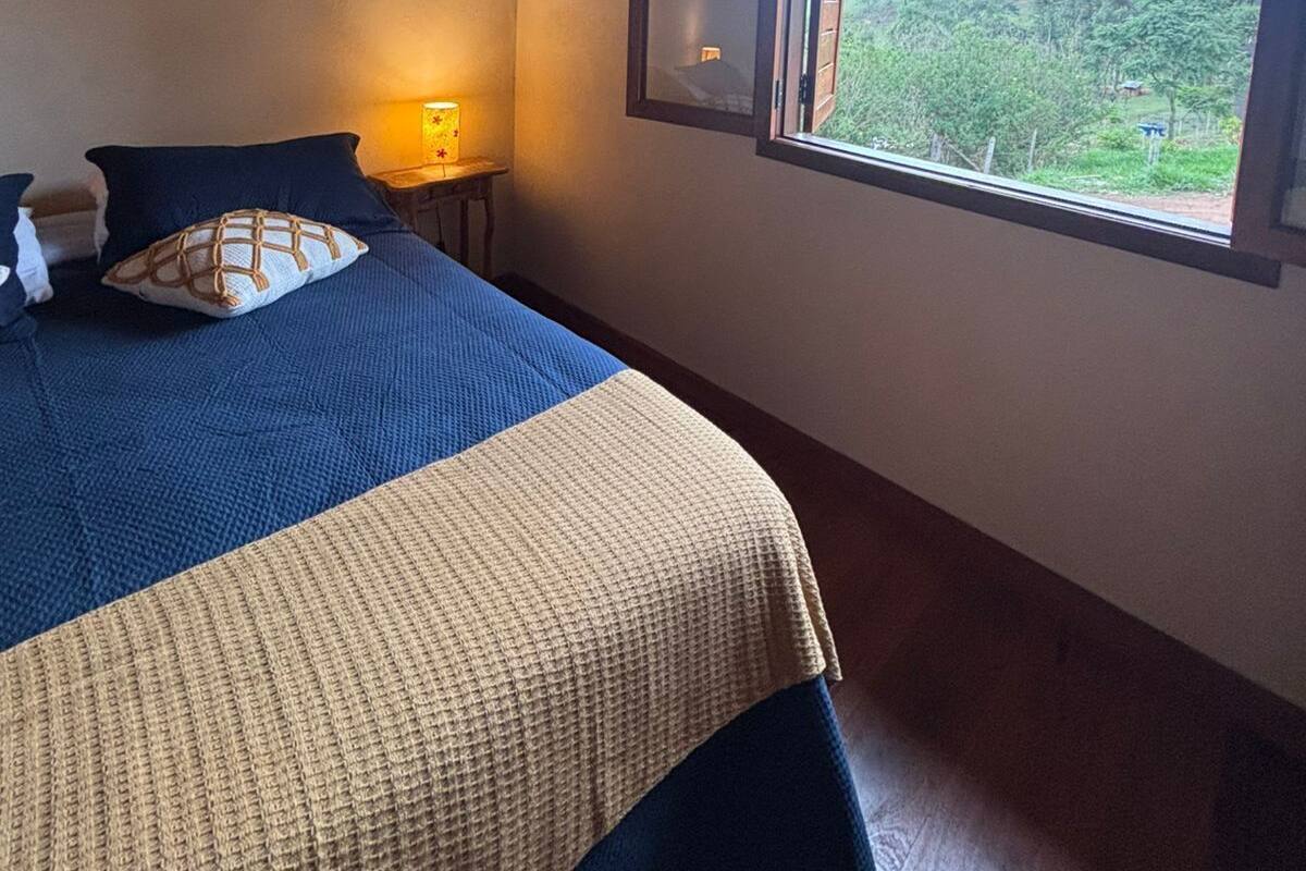 Ein Schlafzimmer mit Holzboden, einem Bett mit blau-beiger Bettdecke, einem Nachttisch mit Lampe und einem Fenster mit Blick auf Grünflächen.