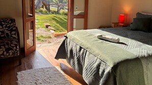 A bedroom with a bed, a chair, a nightstand, a lamp, and a view of a landscape through a door. - Pousada Morada Canto do Sol (São Bento do Sapucaí)