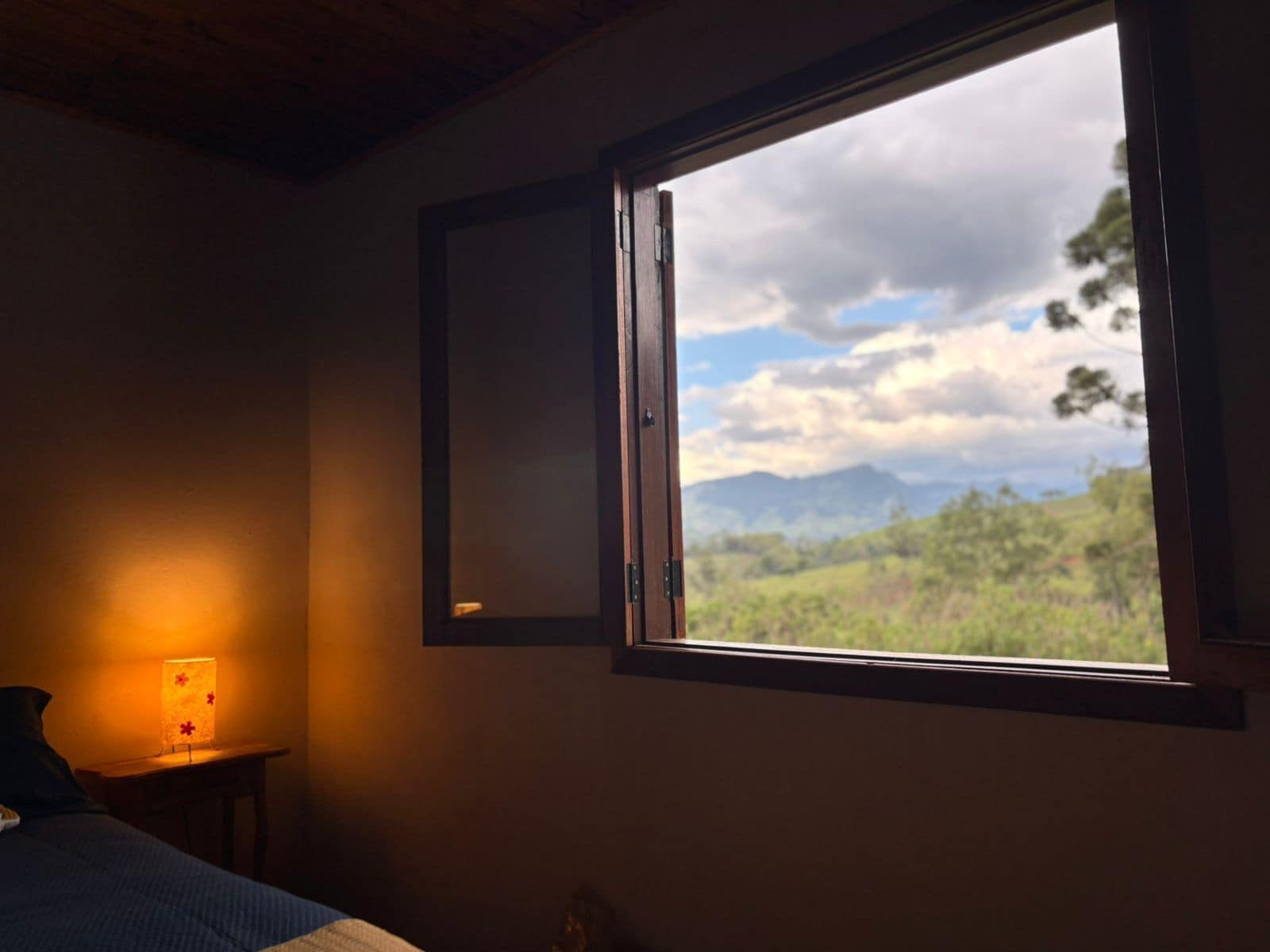 A bedroom with a large window, a bedside lamp, and a view of the outdoors.