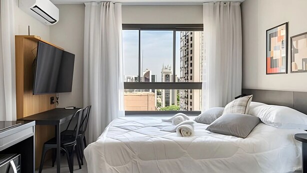 Quarto de hotel com cama, escrivaninha, televisão e janela com vista para a cidade.