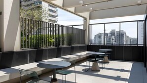 Terraço moderno no telhado com assentos e mesas, grandes janelas com vista para a cidade e uma cerca de metal preto.