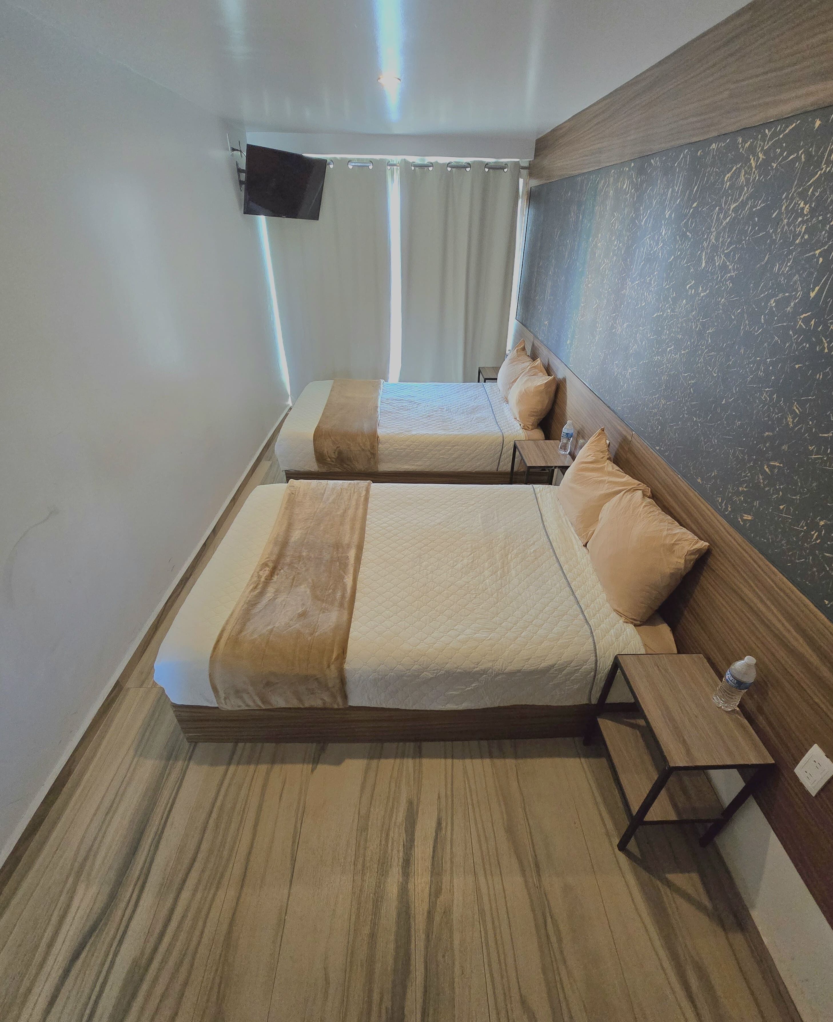 Habitación de hotel con dos camas, un televisor de pantalla plana montado en la pared, una ventana con cortinas y suelo de madera.