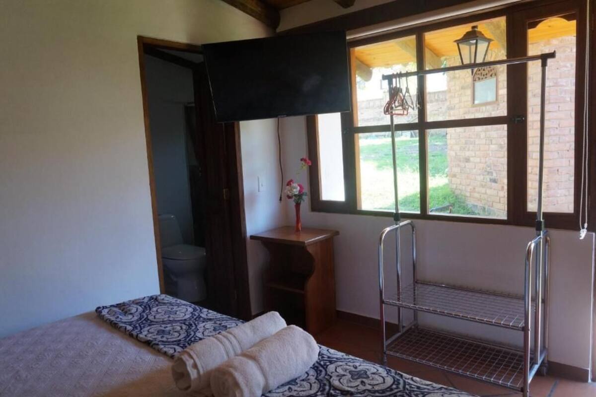 Una habitación con cama, televisión, ventana y vista a un edificio de ladrillo, además de una repisa metálica.