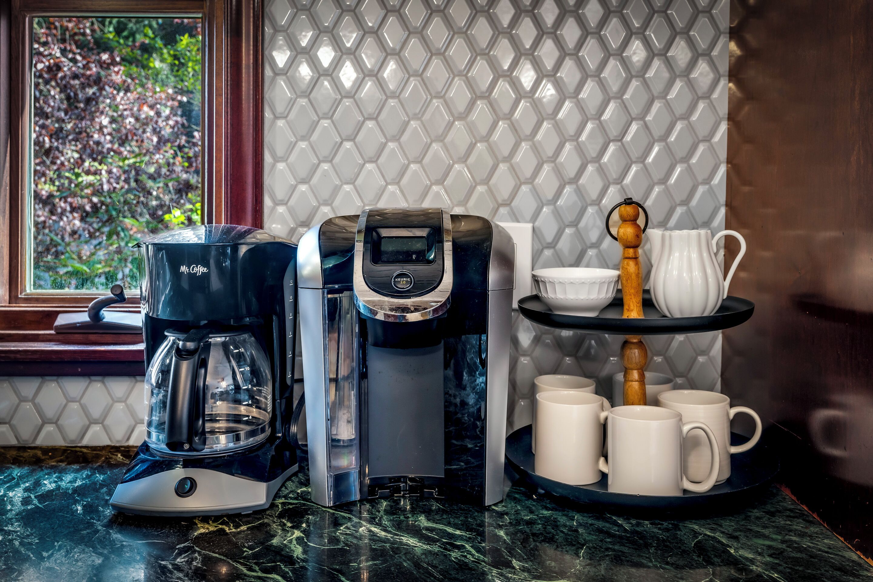 Una cafetera, una jarra de café y una bandeja con tazas y una jarrón en una encimera de cocina.