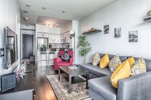 A modern living room with a grey sofa, a red armchair, and a coffee table. There is a kitchen in the background with white cabinets and a microwave. - Amazing Downtown Toronto Condo (Toronto)