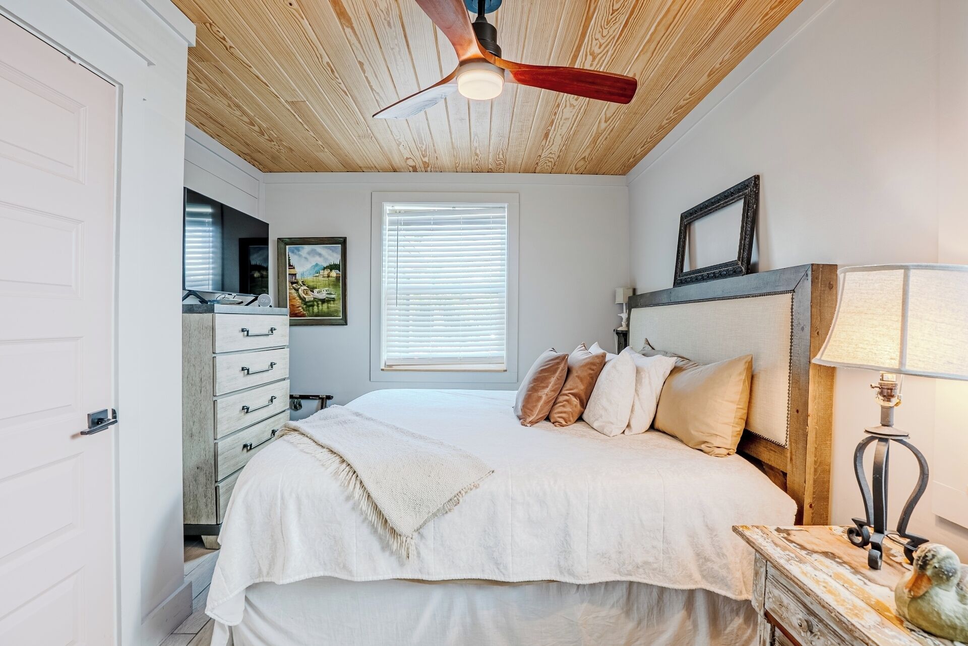 Un dormitorio con cama, ventilador de techo, ventana con persianas, cómoda y lámpara.