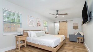 Ein Schlafzimmer mit einem Holzbett, Nachttischen, einem Deckenventilator, einem an der Wand montierten Fernseher und einem Fenster mit Jalousien.
