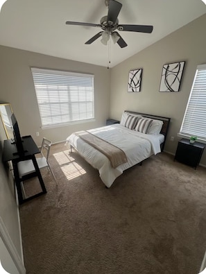 A bedroom with a bed, a desk, a chair, a ceiling fan, and artwork on the walls.