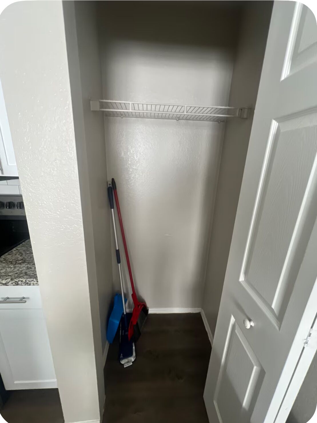 A narrow hallway with a white door on the right, a white shelf above, and a broom leaning against the wall.