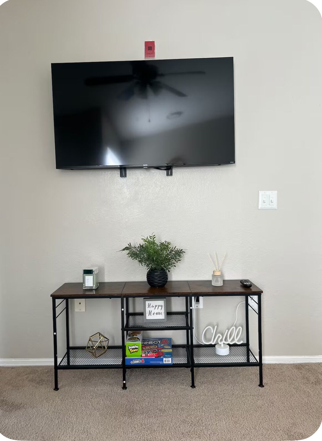 A flat-screen TV mounted on a wall, a dark wooden console table with a plant, a small candle, and a sign that says 'Happy Home.'