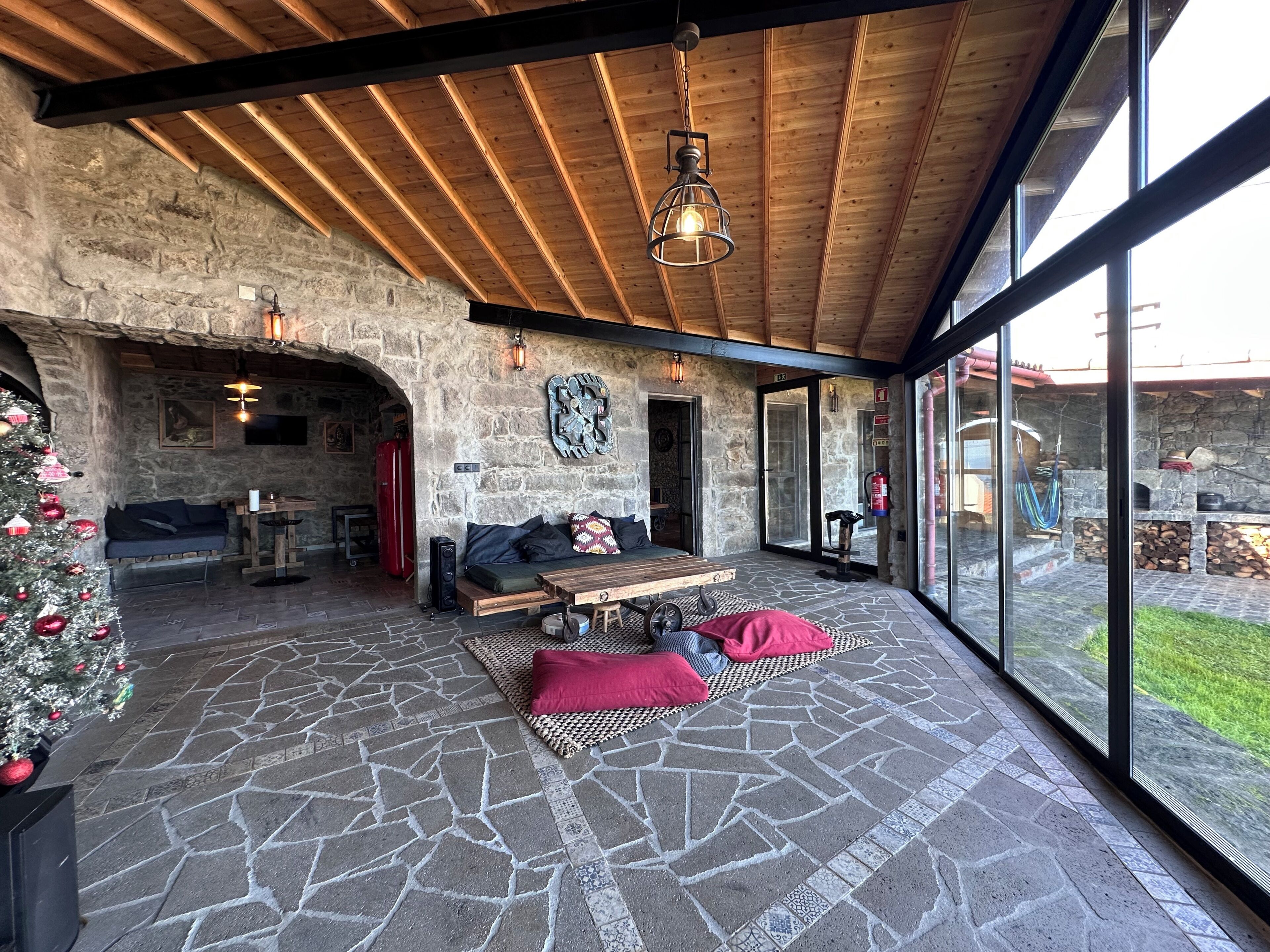 Een interieur met stenen vloer, houten plafond, een zithoek met kussens en uitzicht door grote ramen.