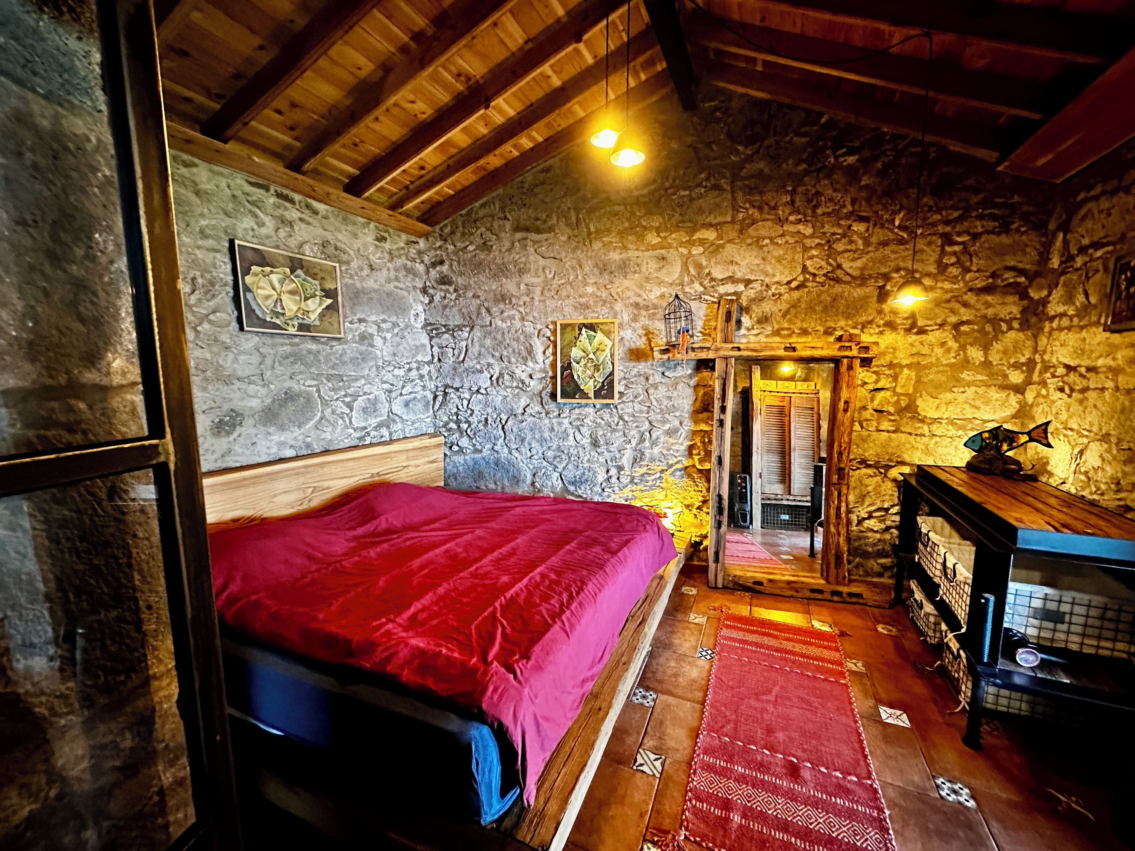 A rustic room with a stone wall, wooden ceiling beams, a bed with a red blanket, a small table with a fish decoration, and a doorway leading to another room.