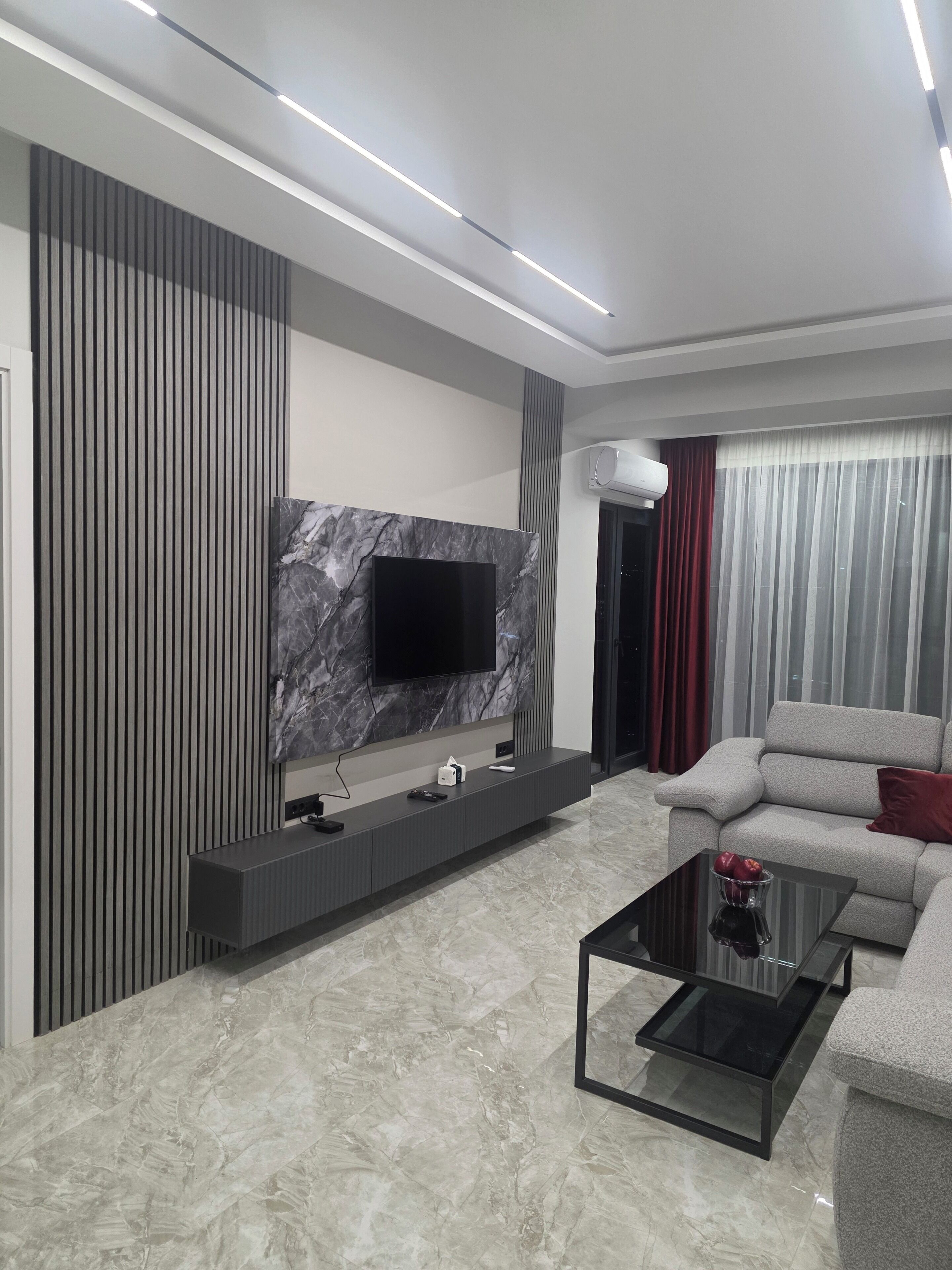 A modern living room with a flat-screen TV mounted on a marble wall, a grey sofa, a glass coffee table, and a built-in entertainment unit.