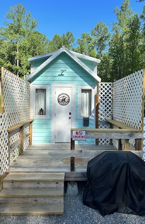 A small, light blue cabin with a white door, a 'NO SMOKING' sign, and a wooden deck leading to it.