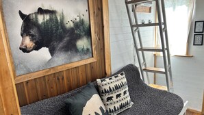 A room with a wooden wall, a framed bear painting, a gray sofa, a ladder, and a window with a view of trees.