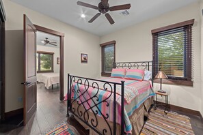 Une chambre à coucher comprenant un lit, un ventilateur de plafond, une fenêtre avec des stores et une porte donnant sur une autre pièce.