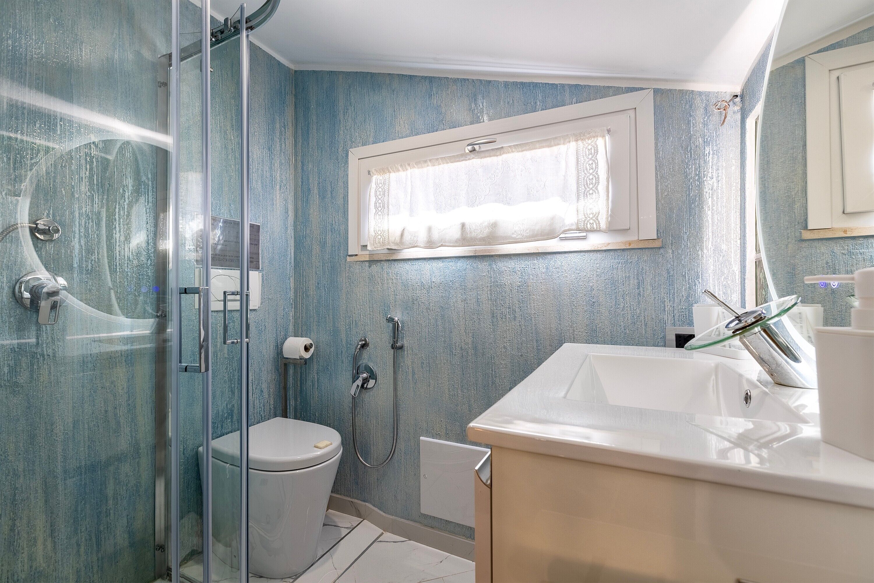 A bathroom with a glass-enclosed shower, a toilet, a sink, and a window with a curtain.