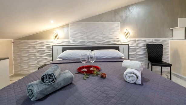 Une chambre d’hôtel moderne avec un grand lit, un plateau rouge avec des verres, des serviettes roulées et une seule rose.