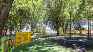 Zona de jocs amb gespa artificial verda, un senyal groc i arbres.
