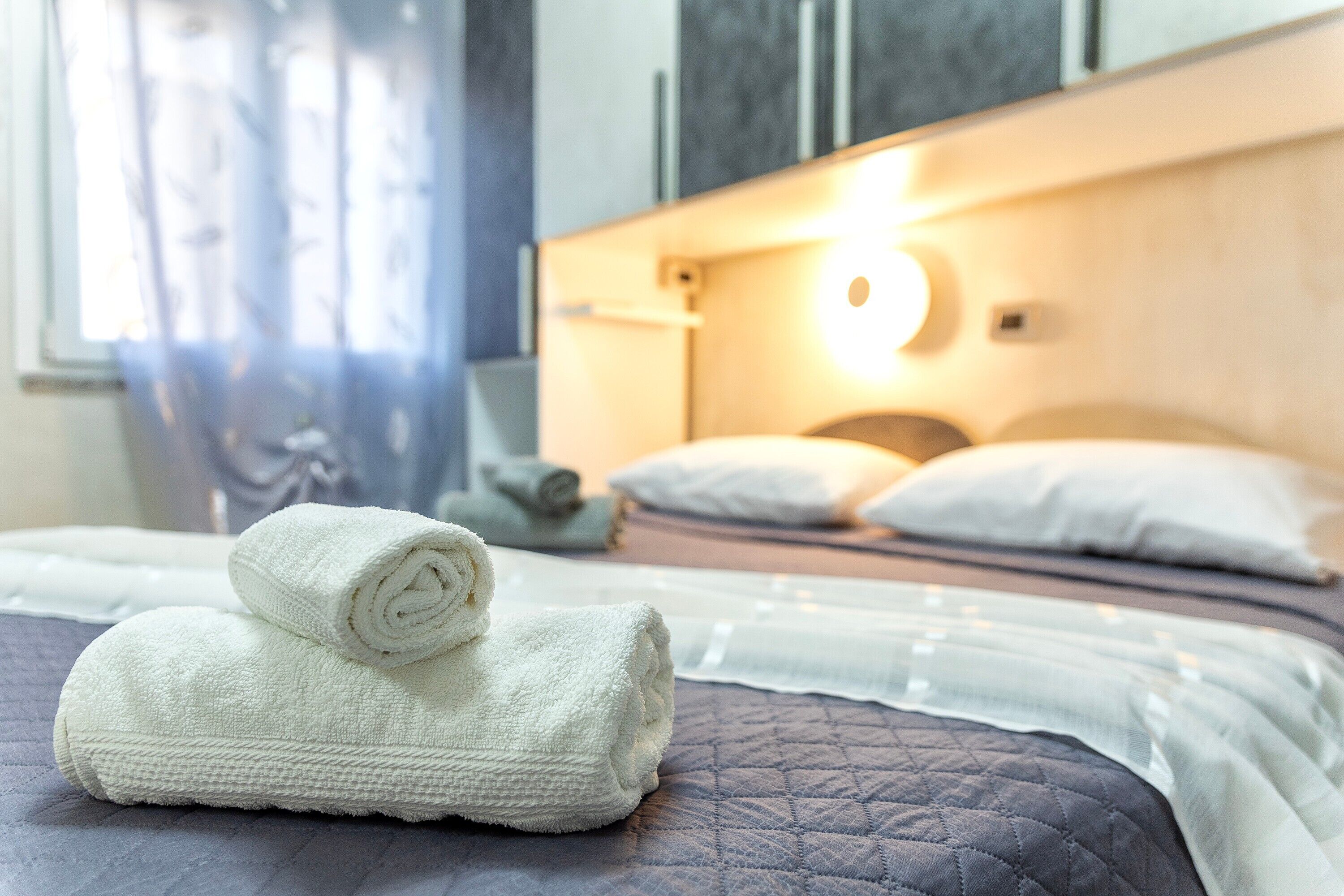 A neatly made bed with a quilted cover, a rolled white towel on the bed, and a bedside lamp.
