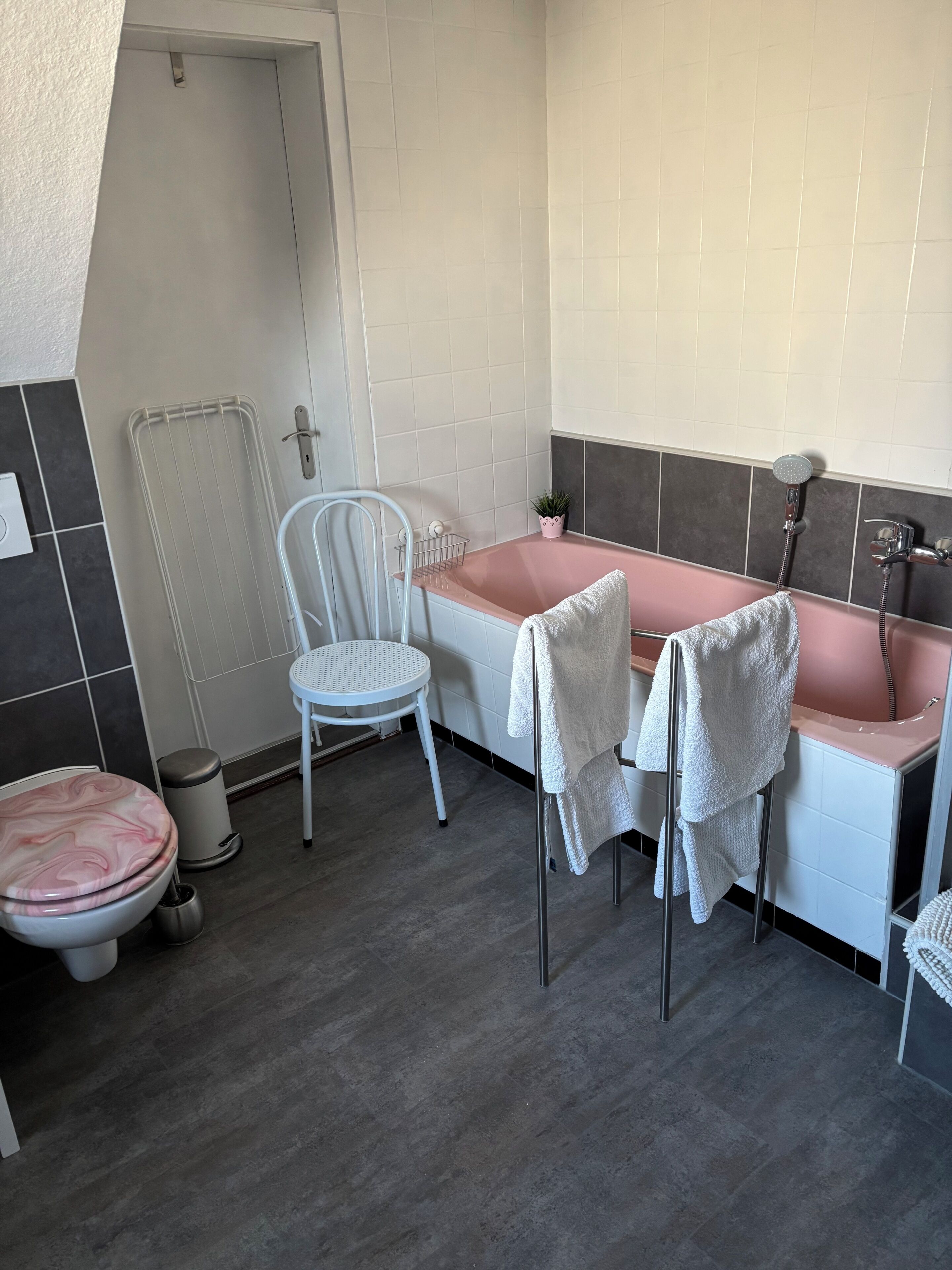 A bathroom with a white toilet, a pink bathtub, a white sink, a white chair, and a small potted plant.