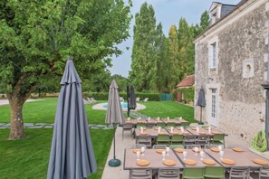 An outdoor dining area with tables and chairs, umbrellas, and a pool in the background. - Villa La Messagerie (49) — Heated pool — Basketball — 2 hours from Nantes — Sleeps 36. (Noyant)