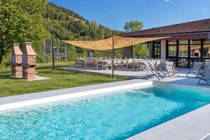 Ein Schwimmbecken mit Liegestühlen und Grillplatz, umgeben von einem angelegten Garten und einem Gebäude mit großen Fenstern.