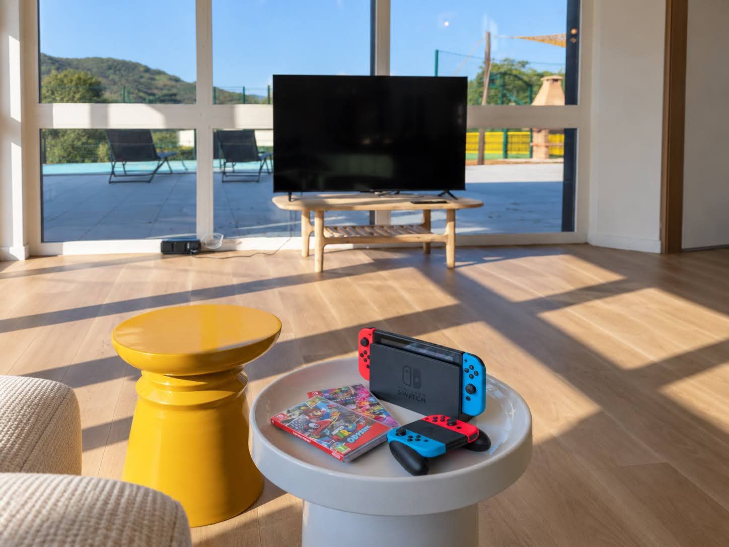 Ein Zimmer mit einem großen Fenster, einem Fernseher, einer Nintendo Switch und einer Zeitschrift auf einem kleinen Tisch.